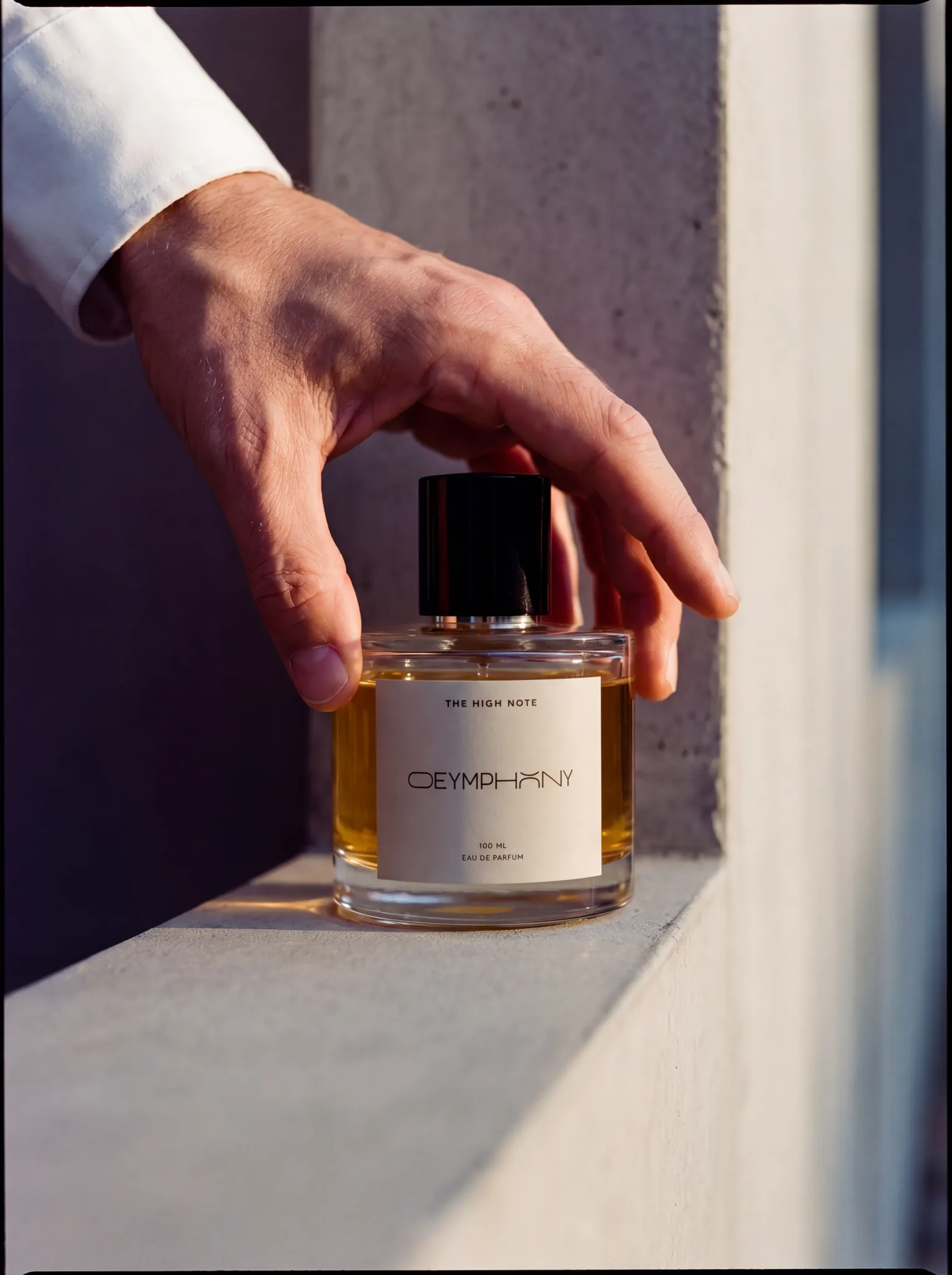 Refined hand draped from above over the cap of The High Note, white cotton cuff visible, smooth concrete ledge, vertical architectural line in soft bokeh.
