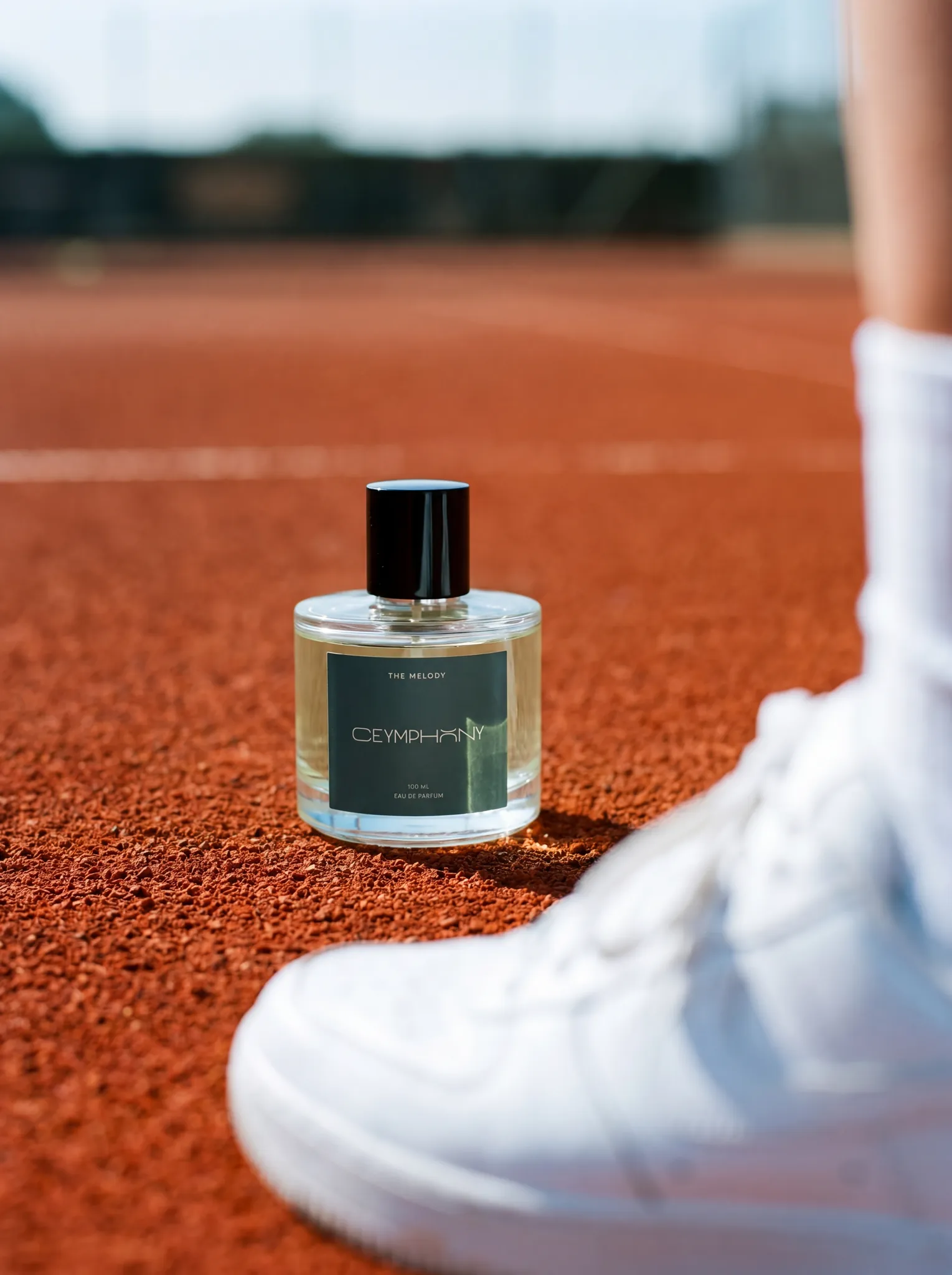 The Melody bottle upright on a red clay tennis court in clean daylight, foreground blur of a white sneaker.