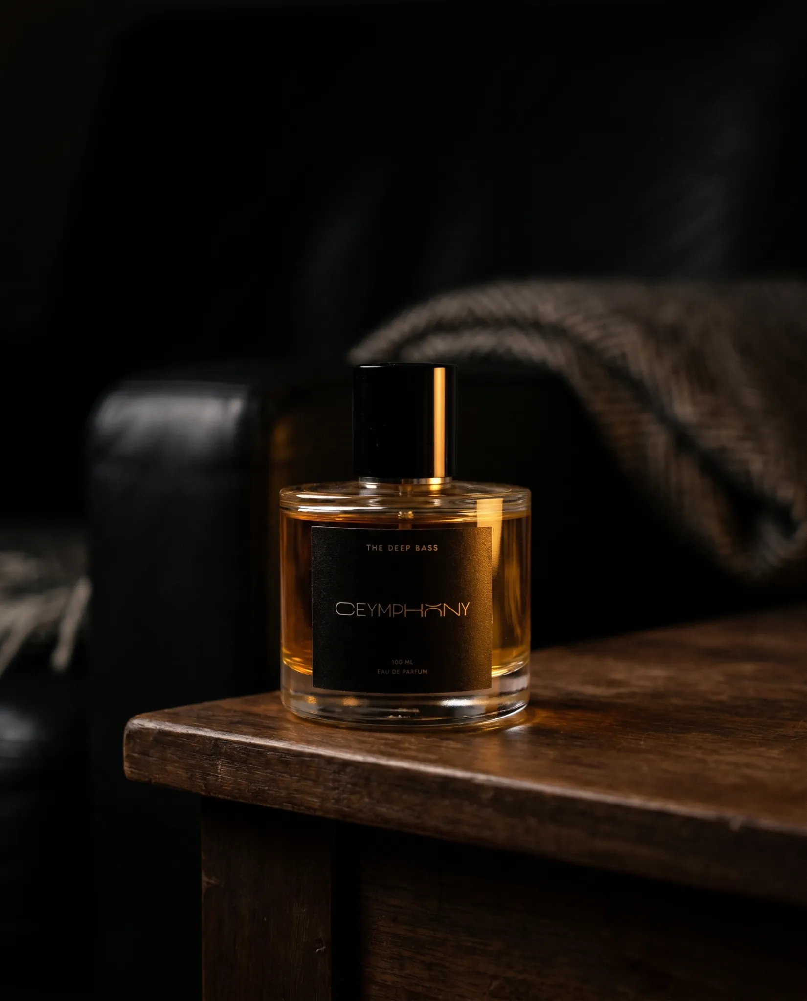 The Deep Bass bottle set down on a dark walnut side table in a dim interior at a late hour, a single tungsten lamp glow on its shoulder, leather chair and wool throw in deep bokeh.